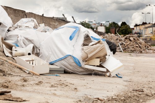 A large pile of assorted construction waste and discarded building materials is situated on a dirt surface at an outdoor site. The debris includes white plastic pipes, broken pieces of drywall or plasterboard, and various discarded packaging materials. Some of the materials are partially covered with a large, crumpled white plastic bag with blue tape or ties. In the background, there is a partially demolished concrete wall with exposed rebar, piles of loose soil, and scattered rubble, indicating ongoing construction or renovation work. Further behind, there are trees, suburban buildings, and streetlights under a cloudy sky, suggesting a suburban area. The scene is typical of a rubbish removal or house clearance operation in the Cobham area, managed by House Clearance Cobham, which handles waste disposal and clearance services for residential clients.