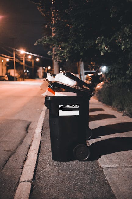 A black wheelie bin with the label 'ST. JOHN'S' positioned on a paved sidewalk beside a curb in a residential area during nighttime. The bin lid is open, revealing a mixture of discarded cardboard, pa