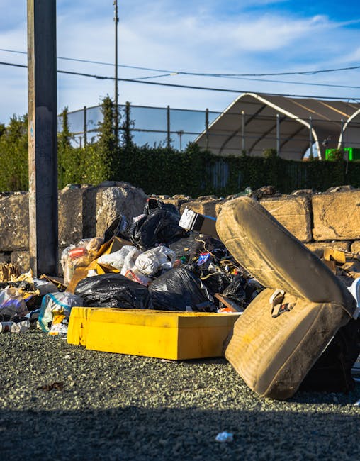 An outdoor scene featuring a large pile of flattened cardboard boxes, primarily cardboard fruit and vegetable trays, stacked on top of each other in a disorganized manner. The boxes are decorated with