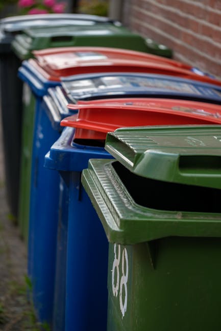 A large, rectangular metal waste bin with a hinged lid, positioned on four black caster wheels on a damp asphalt pathway adjacent to a grassy verge. The bin is weathered, showing signs of dirt and mos