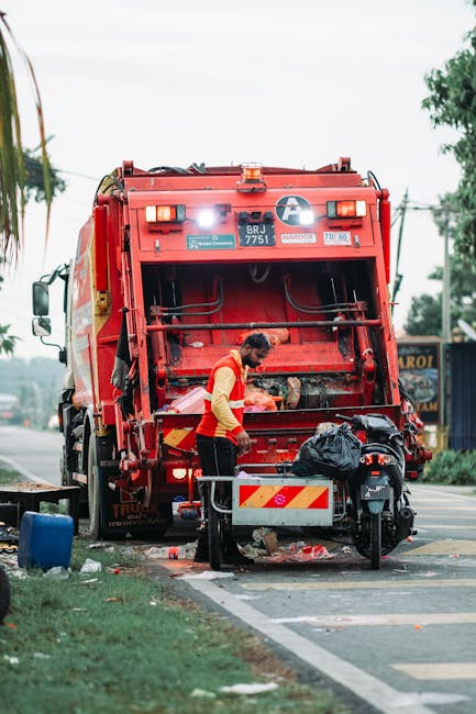A pile of mixed waste items including cardboard boxes, plastic bags, and crumpled packaging materials is situated on the ground next to a brick and concrete wall. The cardboard boxes vary in size and 